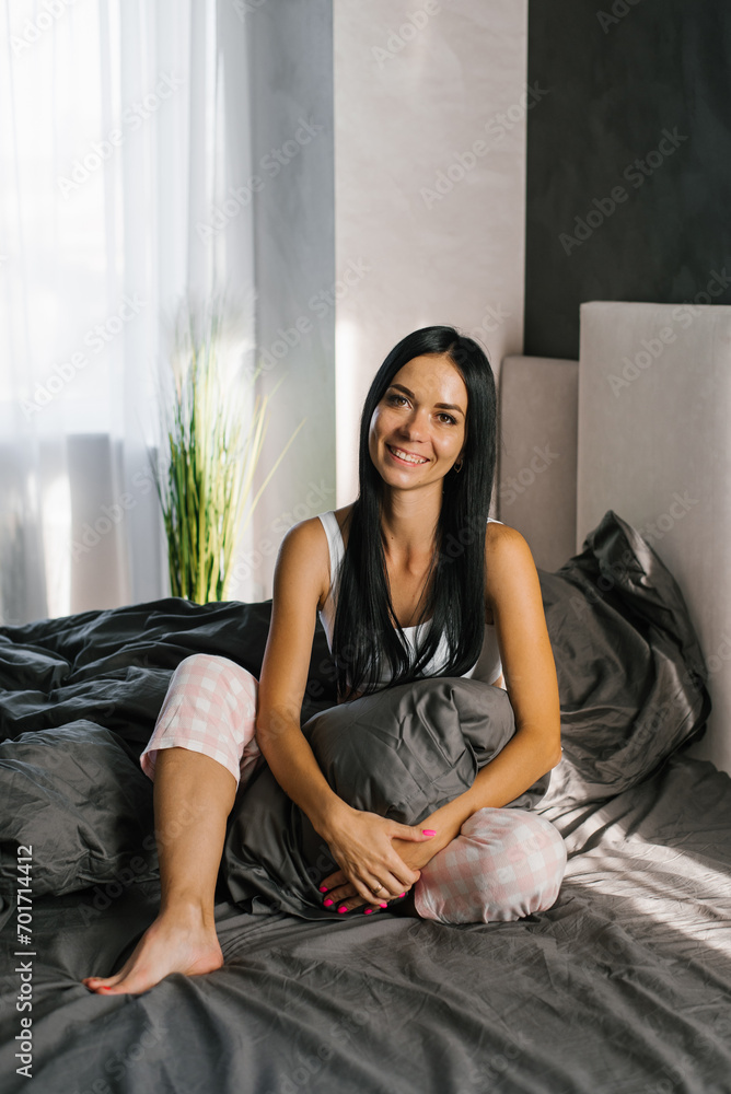 Young Caucasian brunette with long hair in pajamas is sitting on the bed in the morning and smiling Young Caucasian brunette with long hair in pajamas is sitting on the bed in the morning and smiling