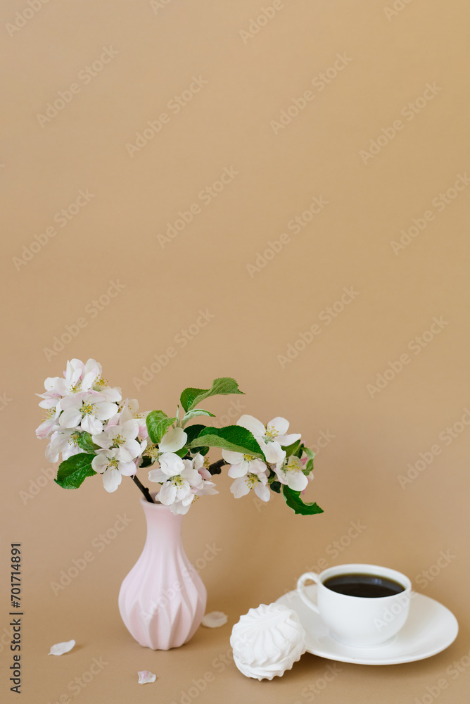 Beautiful branches of a blooming apple tree in a pink vase with a cup of tea and marshmallows on a beige background with a copy space Beautiful branches of a blooming apple tree in a pink vase with a cup of tea and marshmallows on a beige background with a copy space