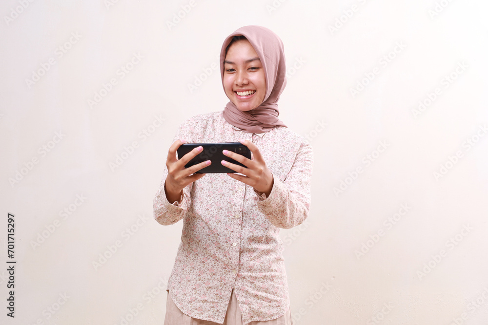 Cheerful young asian muslim girl standing while playing game on her smartphone. Isolated on white background Cheerful young asian muslim girl standing while playing game on her smartphone. Isolated on white background