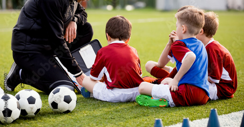 Soccer training for children. Kids and coach sitting on the football field and talking about game strategy. Trainer using tactics board to explain soccer game rules