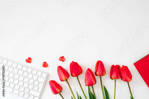 Flat lay of a female office desk with red flowers. Women's workplace with keyboard, tulip flowers, accessories, notepad on a white background. Festive background for Valentine's Day. Copy space #701722603