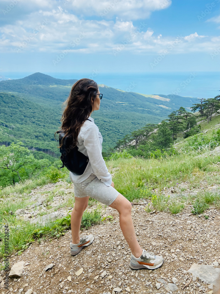 Fototapeta premium Hipster young girl with a backpack enjoying the sunset at the top of the mountain. A tourist traveler on the background of a valley landscape layout. 