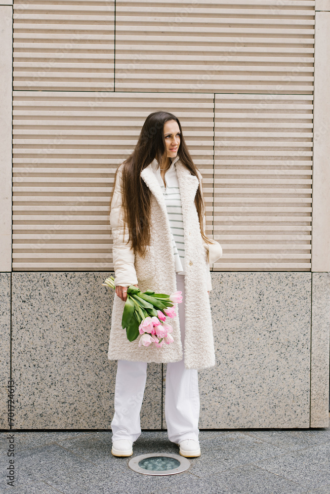 Fototapeta premium Young woman in stylish light coloured clothes strolls through the streets of the city with a bouquet of tulips