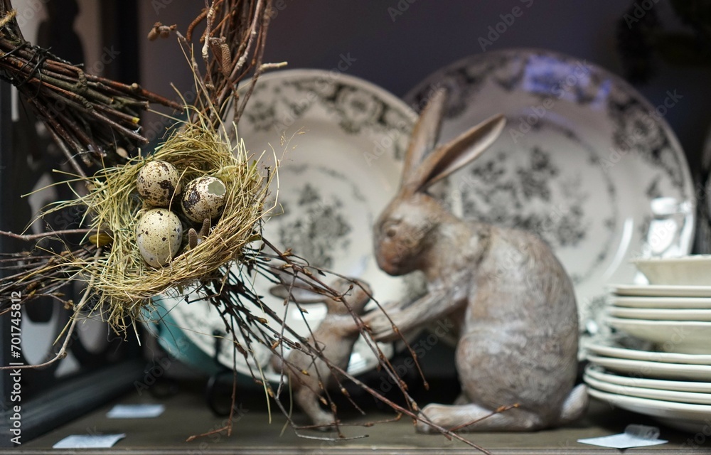  Easter decorations for the house on the shelf in the form of a nest with colorful speckled eggs on the background of a figurine of a gray rabbit