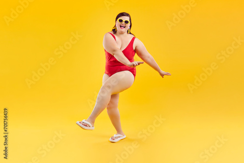 Portrait of overjoyed excited happy funny fat woman in red swimsuit and sunglasses having fun and dancing isolated on a studio yellow background. Summer holiday trip and vacation concept.