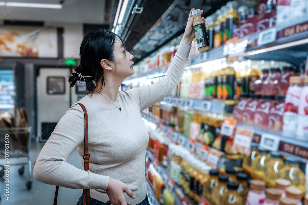 A young Asian girl is shopping in the beverage section of a supermarket, standing by the aisle with a drink reading the label and checking the ingredient list
