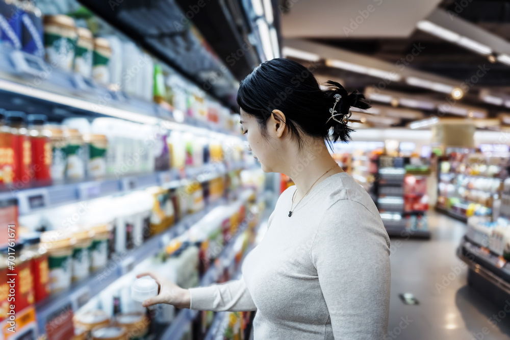 A young Asian girl is shopping in the beverage section of a supermarket, standing by the aisle with a drink reading the label and checking the ingredient list