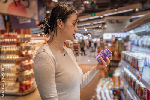 A young Asian girl is shopping in the beverage section of a supermarket, standing by the aisle with a drink reading the label and checking the ingredient list