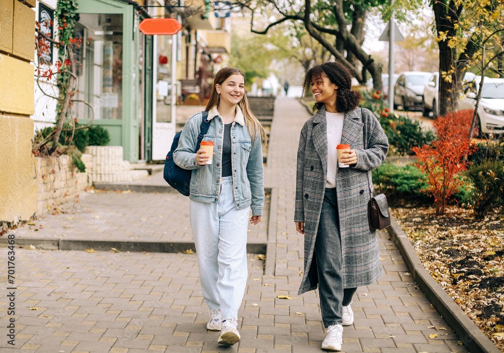 Naklejka premium Two multiracial female best friends walking city street with take away coffee, enjoying autumn walk