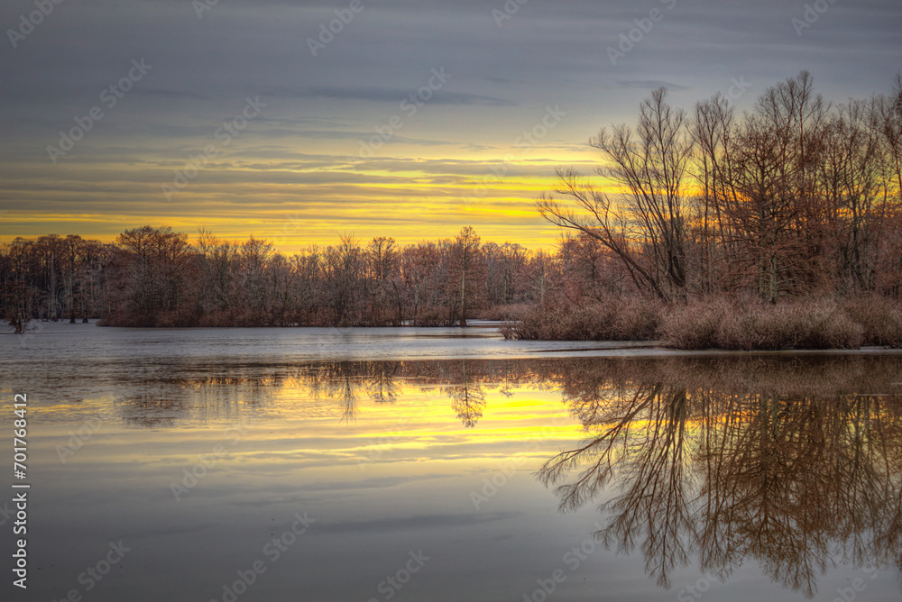 A winter evening on Horseshoe Lake. Patches of thin ice on the surface.  Bare trees on the horizon.  Sky in layers of colors.  Reflections where the ice has melted. 