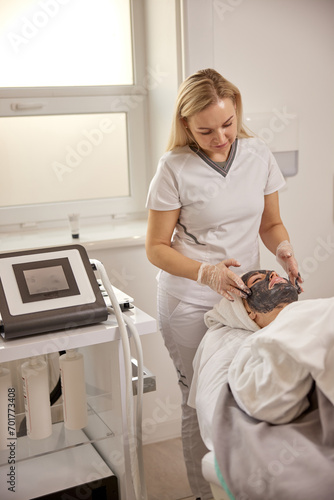 Well-lit spa, Beauty spa, Skin rejuvenation. Esthetician carefully covers client's face with detoxifying charcoal mask, aiming for comprehensive cleanse in well-lit confines of spa.