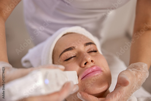 Ultrasound facial, Skincare routine, Beauty enhancement. Concentrated beautician performing ultrasound facial therapy on client, focusing on skin rejuvenation and care in clinical beauty setting.