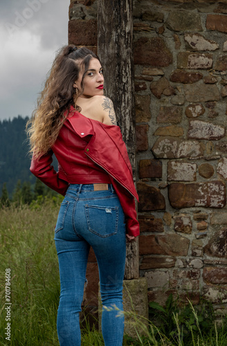 young woman in jeans and a red jacket stands by a stone structure amidst greenery under a cloudy sky