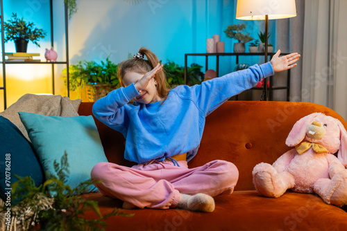 Trendy cheerful positive redhead girl having fun dancing, moving to rhythm, dabbing raising hands, making dub dance gesture. Teenager child kid at home in night evening play room sitting on couch