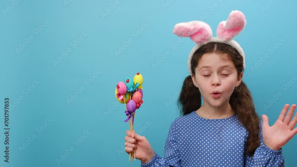Portrait of shocked happy little girl kid wearing pink bunny ears holding colored eggs for Easter, looking excitedly at camera, posing isolated on blue color background wall in studio. Holiday concept