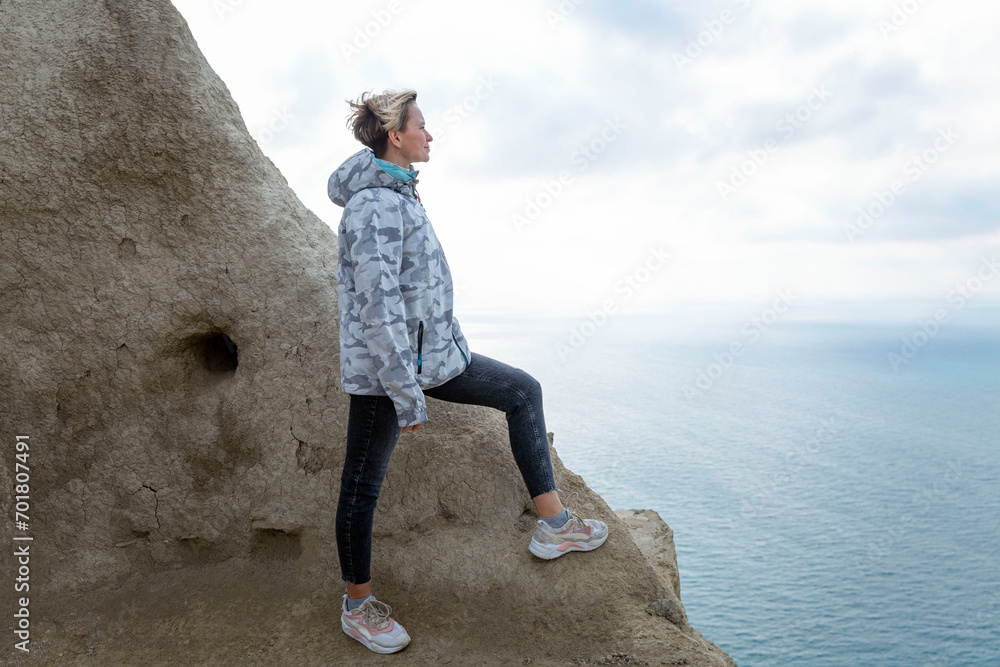 A woman looks down at the sea while standing on a mountain. Cute blonde in a gray jacket. Active recreation in nature.