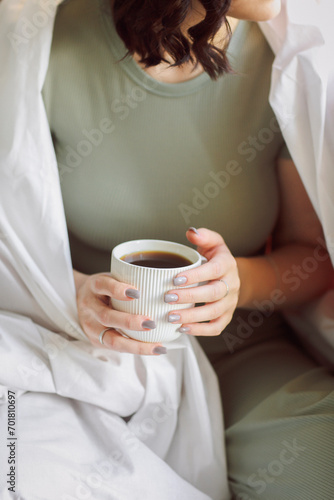 Relaxed positive young brunette woman in homewear sitting on top of unmade bed with cup of tea in her hands, slowly drinking aromatic hot beverage while enjoying spending free time alone at home. 