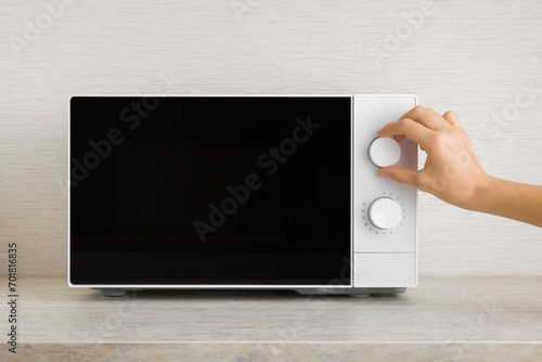 Young adult woman hand rotating white button of dark black microwave oven and setting temperature or functions for food warming on table top at home kitchen. Closeup. Front view. photo