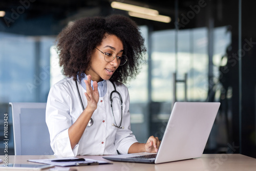African american young female doctor sitting in clinic office at table and talking online with patient, consulting and greeting with hand to camera