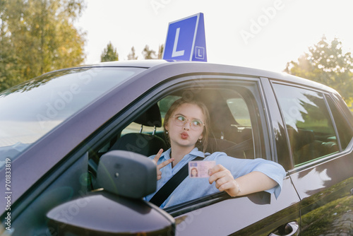 Young beautiful woman successfully passed driving school test. Female smiling and holding driver's license in a brown car with blue L plate on a roof. © Iryna