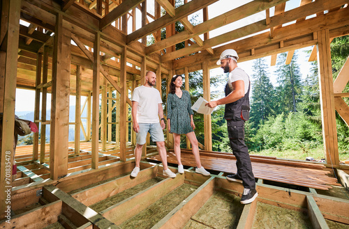 Man worker with detailed plan in hand showcases to young couple process of constructing wooden framed house. Youthful pair of investors inspecting their future abode in mountains near forest.