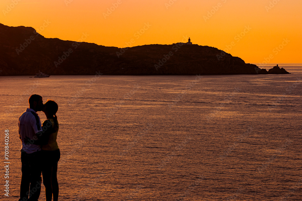 Couple of lovers kissing while posing in front of Revellata Lighthouse, Calvi bay in Corsica, at sunset. Young beautiful couple in love posing in front of sunset.