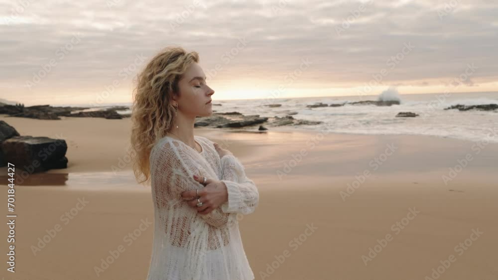 A young woman in white stands alone on a beach, hugging herself, and contemplating the ocean at sunset A young woman in white stands alone on a beach, hugging herself, and contemplating the ocean at sunset