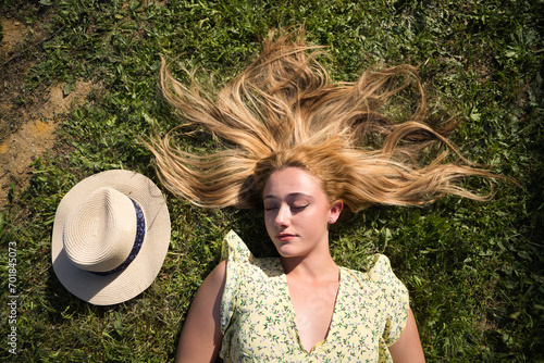Portrait of a blonde woman with long hair, young and beautiful, lying on fresh green grass. Next to her a straw hat. The girl is happy and smiling. Concept holidays and free time. #701845073