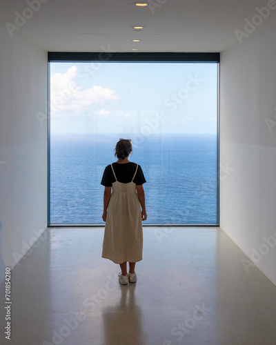 Frau blickt mit Sehnsucht durch modernes Panoramafenster aufs weite Meer
