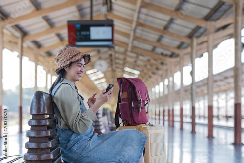 Young Asian woman in modern train station Female backpacker passenger sitting on a bench using a phone while waiting for a train at the train station to go on holiday.