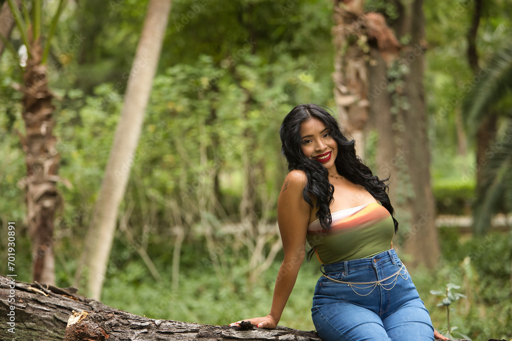 Naklejka premium South American woman, young, beautiful, brunette, with colorful top and jeans sitting on a tree trunk, smiling, happy and relaxed. Concept of beauty, fashion, trend, ethnicity, diversity.