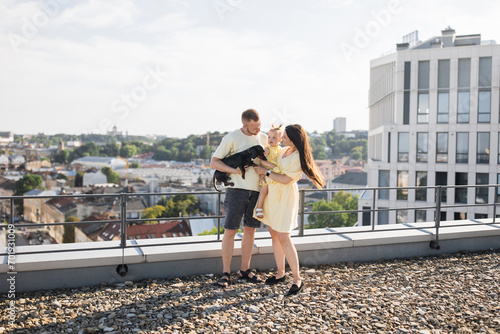Pleased parents in summer clothes standing together on roof of modern city building. Nice young family having memorable moments while holding their cute baby-girl and dog in their arms.