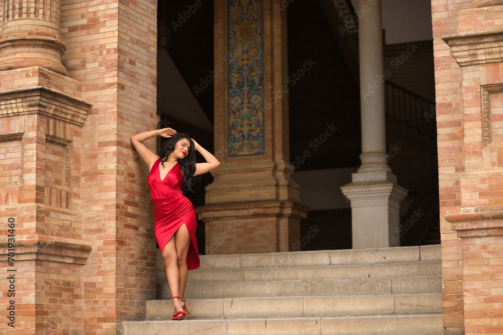 South American woman, young, beautiful, brunette, with an elegant red dress, posing as a model in the square of Spain in Seville. Concept of beauty, fashion, trend, ethnicity, diversity, elegance.