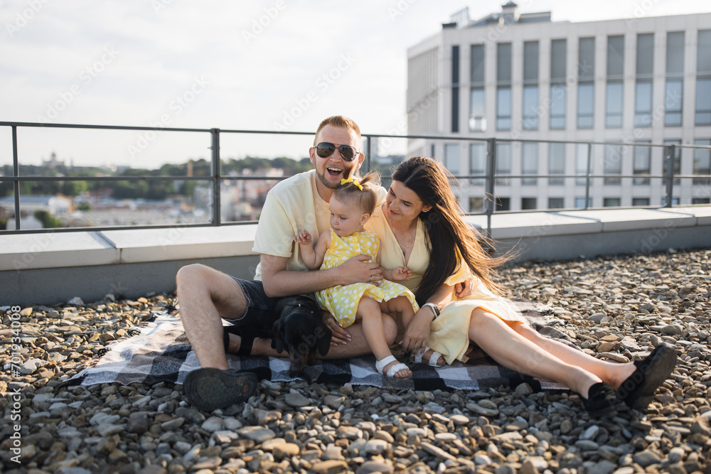 Joyful adult father smiling and sitting near wife while holding small daughter and dog on lap. Caucasian stylish spouses having fun together and giving kid happy childhood on roof of building.