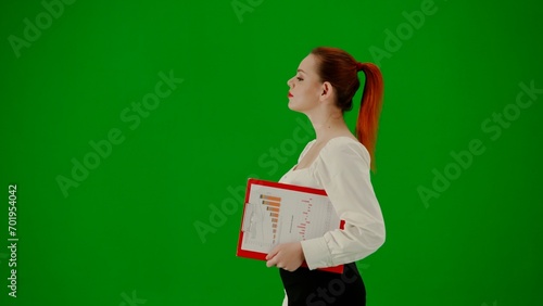 Portrait of attractive office girl on chroma key green screen. Woman in skirt and blouse walking holding red folder. Side view.