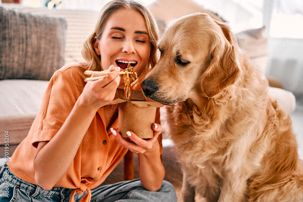 Obraz na płótnie Togetherness with animal. Golden retriever eagerly licking paper box with chinese noodles being eating by young woman. Female pet owner sharing food with lovely furry friend at cozy apartment. w salonie