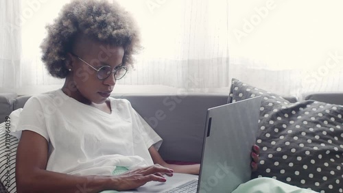 African American woman working with a laptop sitting on a sofa in an apartment. Work at home photo