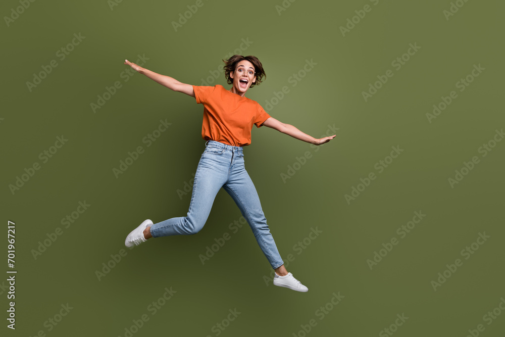 Full size photo of funky woman dressed orange t-shirt jeans in glasses flying hold arms like wings isolated on khaki color background Full size photo of funky woman dressed orange t-shirt jeans in glasses flying hold arms like wings isolated on khaki color background