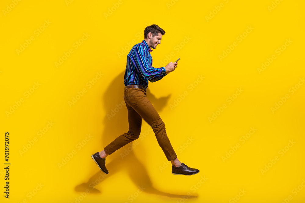 Side profile full size photo of busy guy dressed striped shirt brown trousers run look at smartphone isolated on yellow color background
