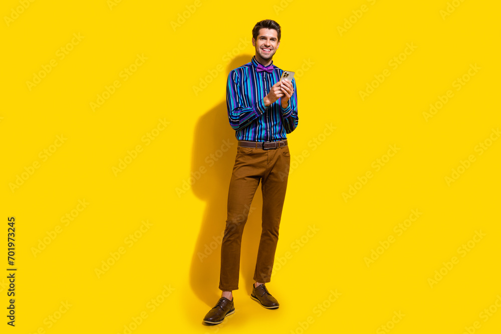 Full size photo of intelligent guy dressed striped shirt trousers hold smartphone read notification isolated on yellow color background Full size photo of intelligent guy dressed striped shirt trousers hold smartphone read notification isolated on yellow color background