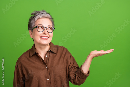 Portrait of pleasant person with gray hairdo dressed brown blouse look empty space palm hold product isolated on green color background