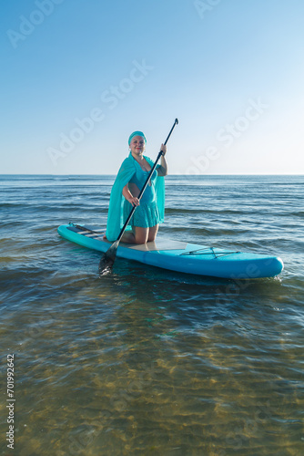 A woman in a turquoise swimsuit with a skirt and a scarf on her head on a SUP board near the sea © finist_4