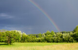 Real rainbow in dark sky. Countryside landscape with fields and trees.