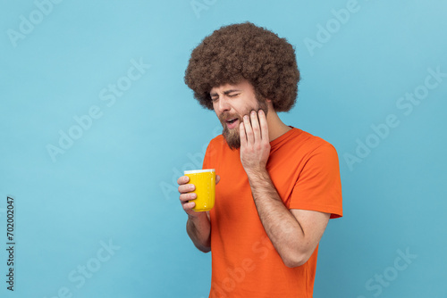 Portrait of man with Afro hairstyle wearing orange T-shirt suffering from terrible teeth pain after drinking hot or cold beverage, dental injury. Indoor studio shot isolated on blue background.
