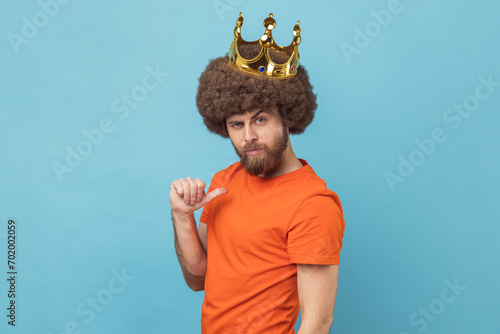 Portrait of serious man with Afro hairstyle wearing orange T-shirt pointing himself, looking at camera with smile, superior privileged status. Indoor studio shot isolated on blue background.