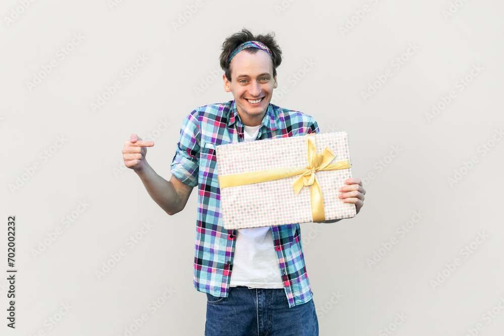 Portrait of attractive smiling positive man holding and pointing at present box, expressing happiness, wearing blue checkered shirt and headband. Indoor studio shot isolated on gray background. Portrait of attractive smiling positive man holding and pointing at present box, expressing happiness, wearing blue checkered shirt and headband. Indoor studio shot isolated on gray background.