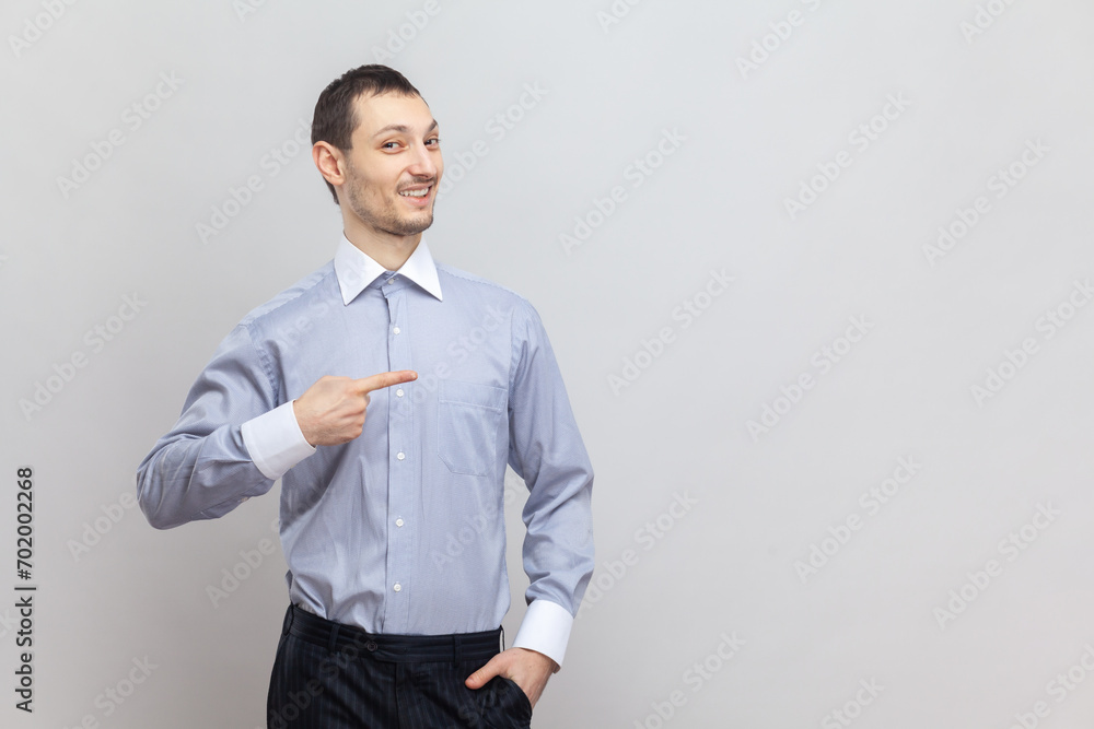Portrait of smiling positive optimistic man standing pointing aside and advertisement area, showing copy space for promotion, wearing light blue shirt. Indoor studio shot isolated on gray background. Portrait of smiling positive optimistic man standing pointing aside and advertisement area, showing copy space for promotion, wearing light blue shirt. Indoor studio shot isolated on gray background.