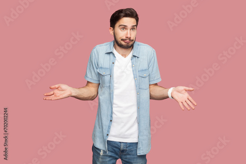 Portrait of unaware questioned man standing spreads palms, shrugs shoulders with perplexed expression, being indecisive, has no idea what happened. Indoor studio shot isolated on pink background.