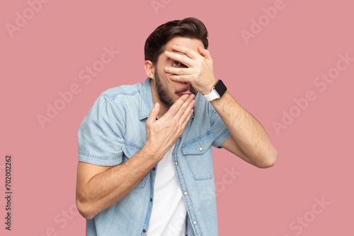 Portrait of scared frightened bearded man in blue casual style shirt standing peeks through fingers, hides face, stares with at something spooky. Indoor studio shot isolated on pink background. photo