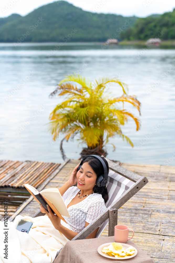Asian woman sitting on lake house balcony reading a book and listening to the music on headphones with mobile app. Attractive girl enjoy outdoor lifestyle travel nature on summer holiday vacation.